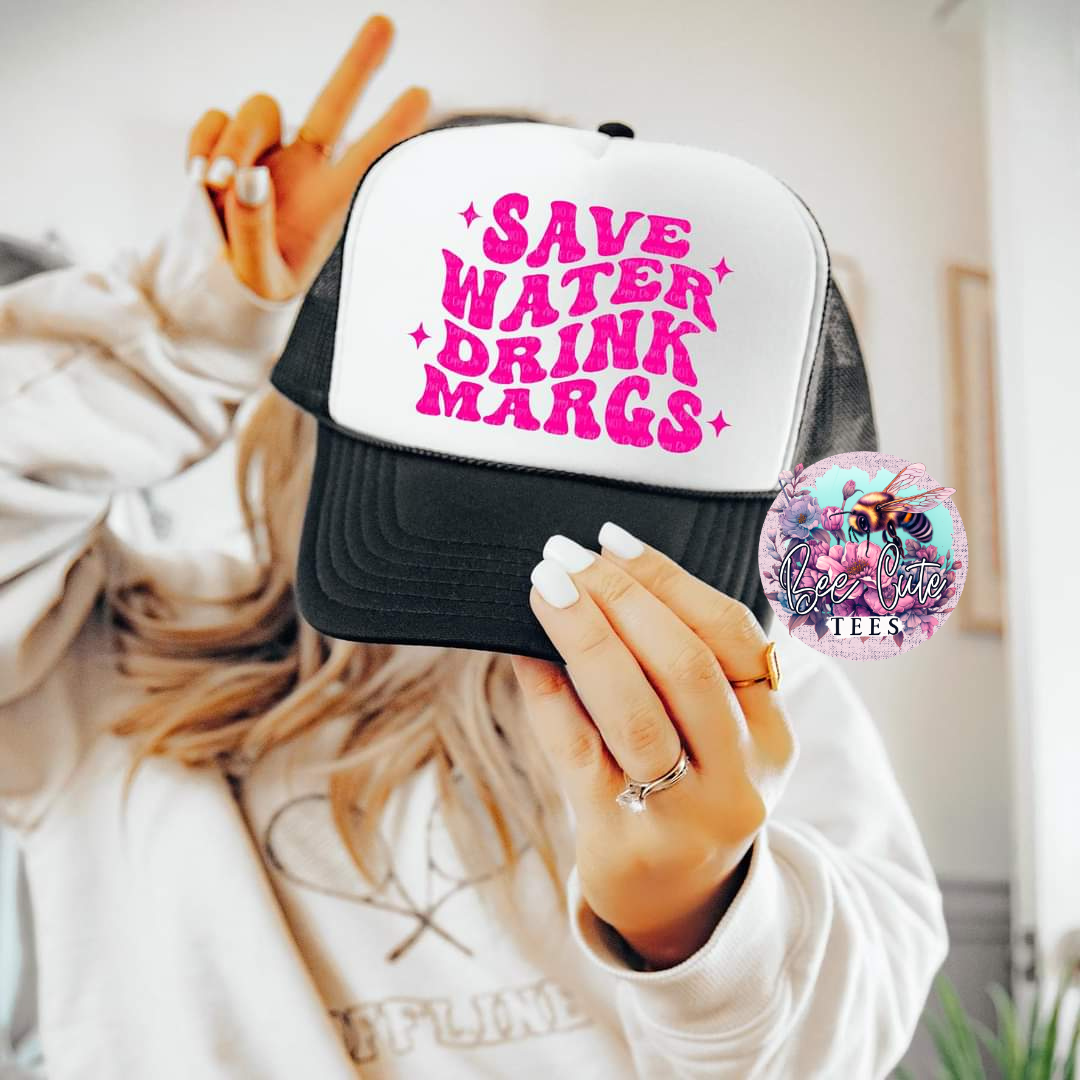 Black and white trucker hat with bold pink “Save Water Drink Margs” graphic, held up by a woman in a casual sweatshirt making a peace sign, for a fun and playful vibe.
