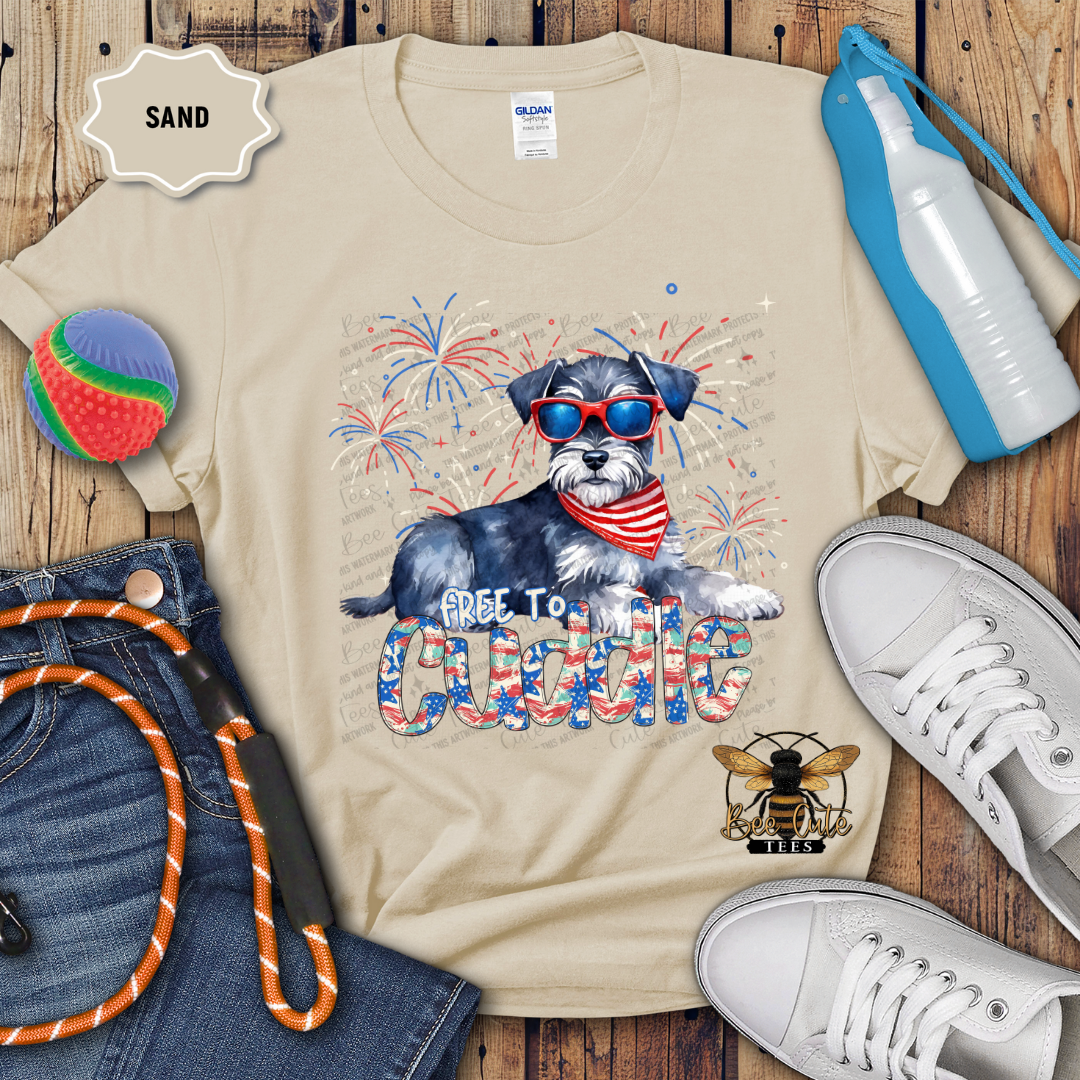 A beige colored t-shirt with a graphic print of a Schnauzer dog wearing sunglasses and a 'Free To Cuddle' text, surrounded by Fourth of July themed decorations, including a red, white, and blue water bottle, a patriotic pin, and a pair of shoes.