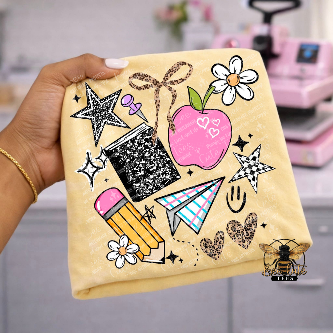 A hand holds a folded  yellow t-shirt, decorated with a playful school-themed graphic print. The print features a pink apple with hearts, a black glittery notebook, a yellow pencil with a pink eraser, a blue-and-white striped paper airplane, white daisy flowers, and leopard-print hearts.