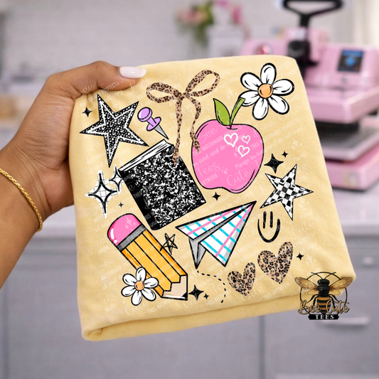 A hand holds a folded  yellow t-shirt, decorated with a playful school-themed graphic print. The print features a pink apple with hearts, a black glittery notebook, a yellow pencil with a pink eraser, a blue-and-white striped paper airplane, white daisy flowers, and leopard-print hearts.