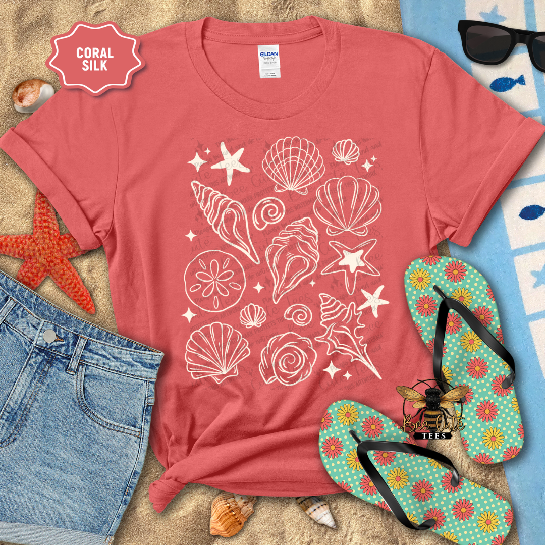 Coral t-shirt with seashell design on a beach-themed background with sunglasses, flip-flops, and starfish.