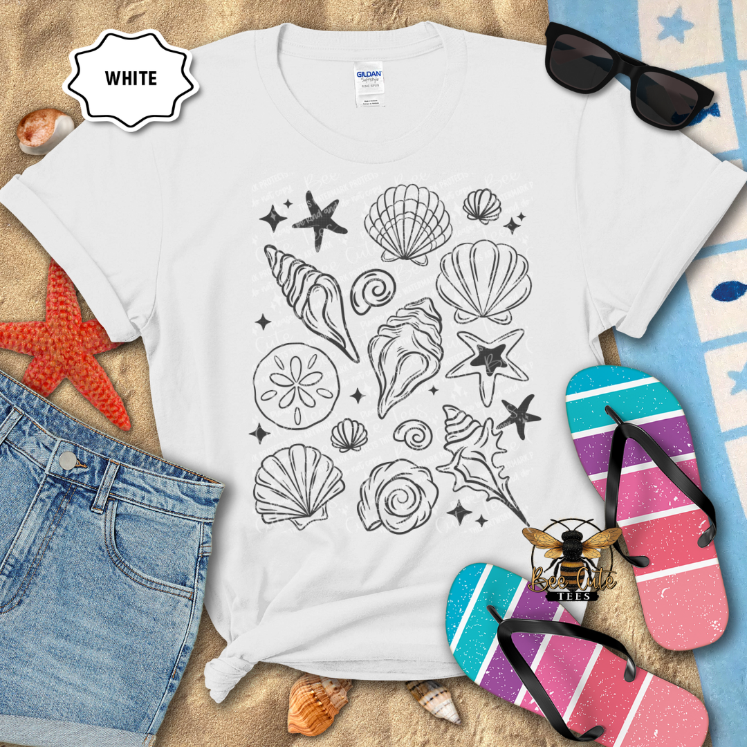White t-shirt with black sea-themed design on a sandy surface with beach accessories.