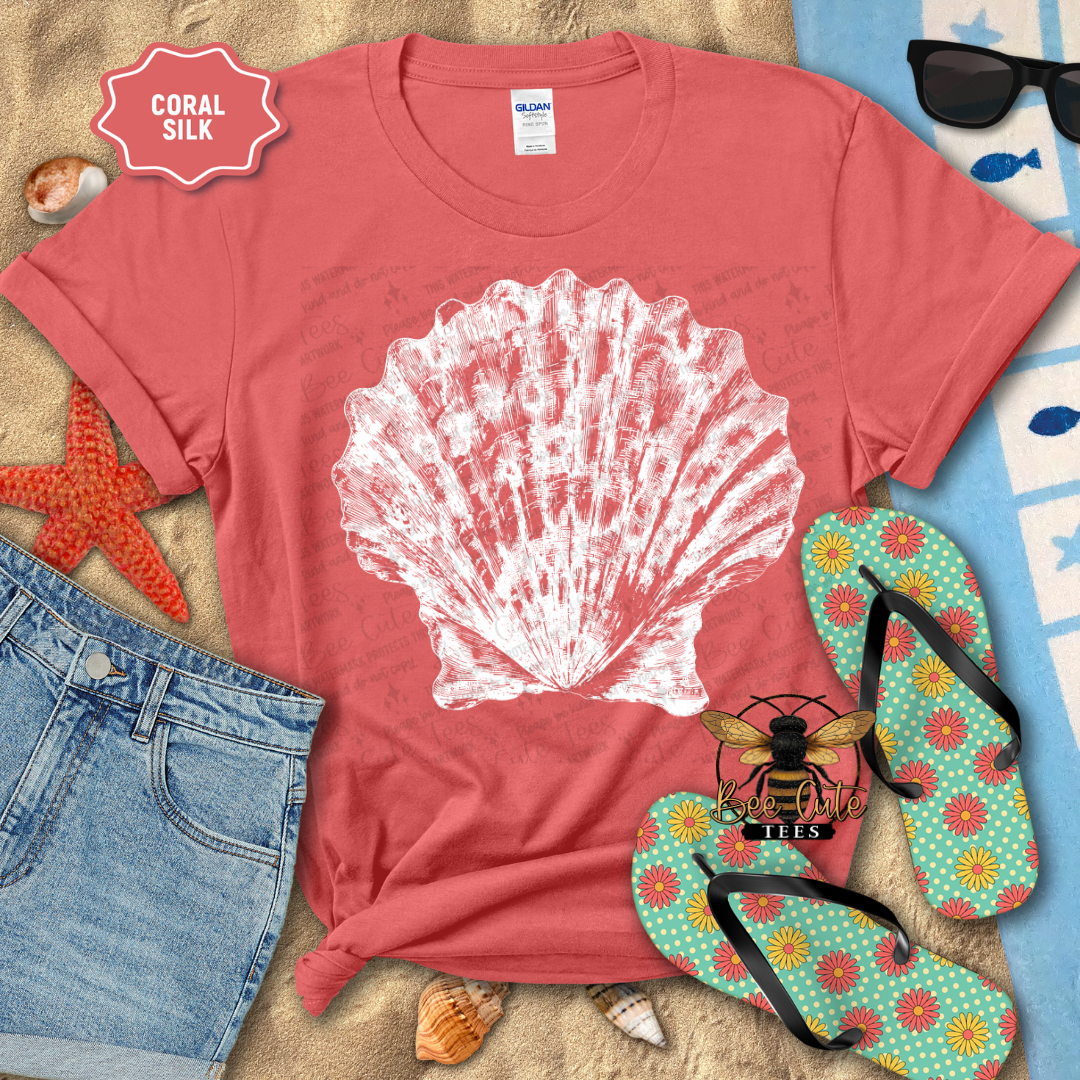 Coral t-shirt with a white shell design, surrounded by beach accessories on sand.