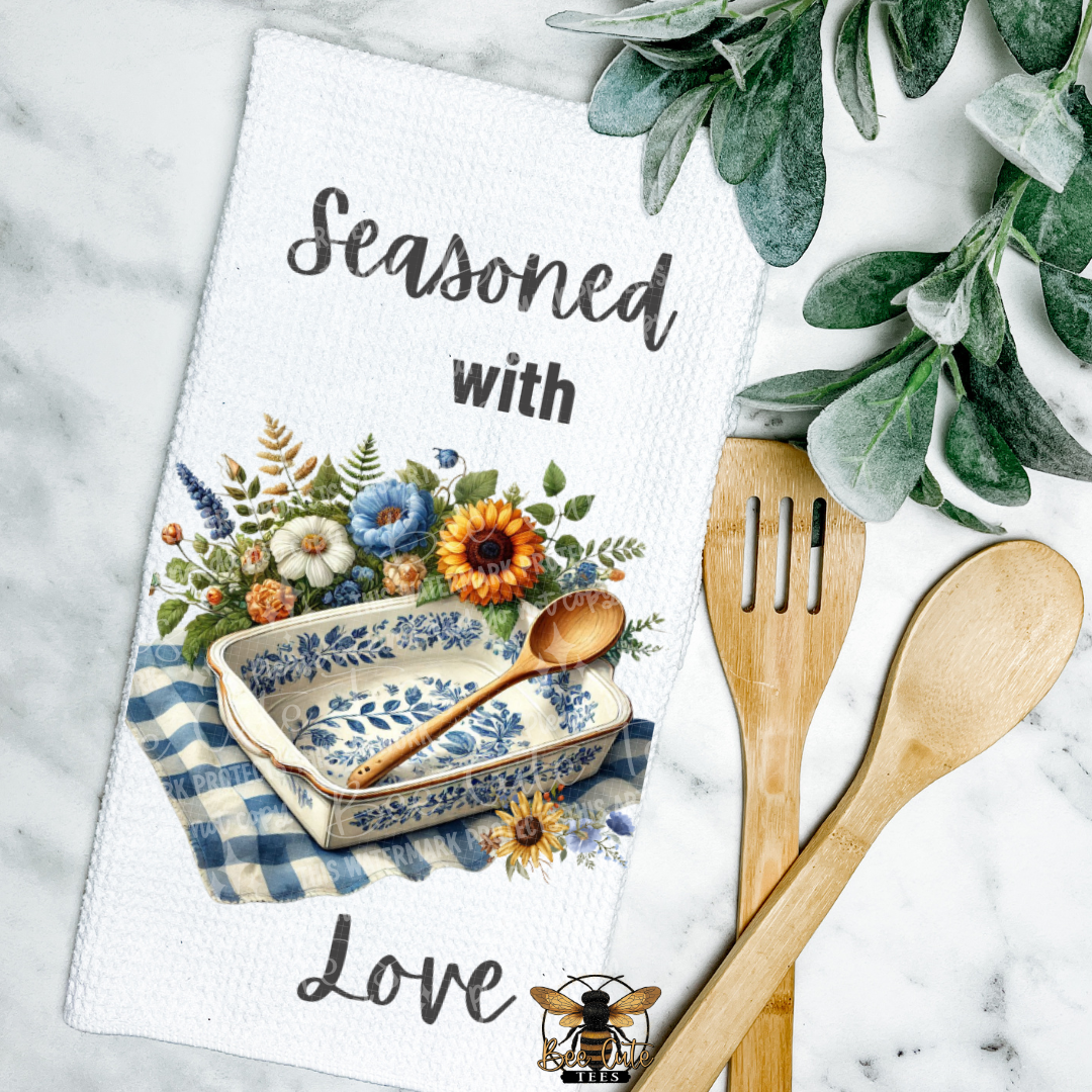 Kitchen towel with floral design and text 'Seasoned with Love' on a marble surface with wooden utensils and greenery.