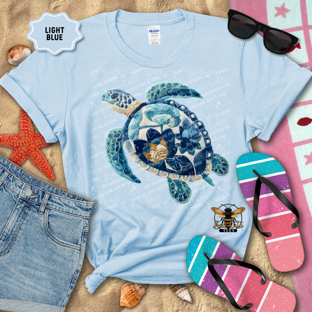 Light blue t-shirt with a turtle design on a sandy background with beach accessories.