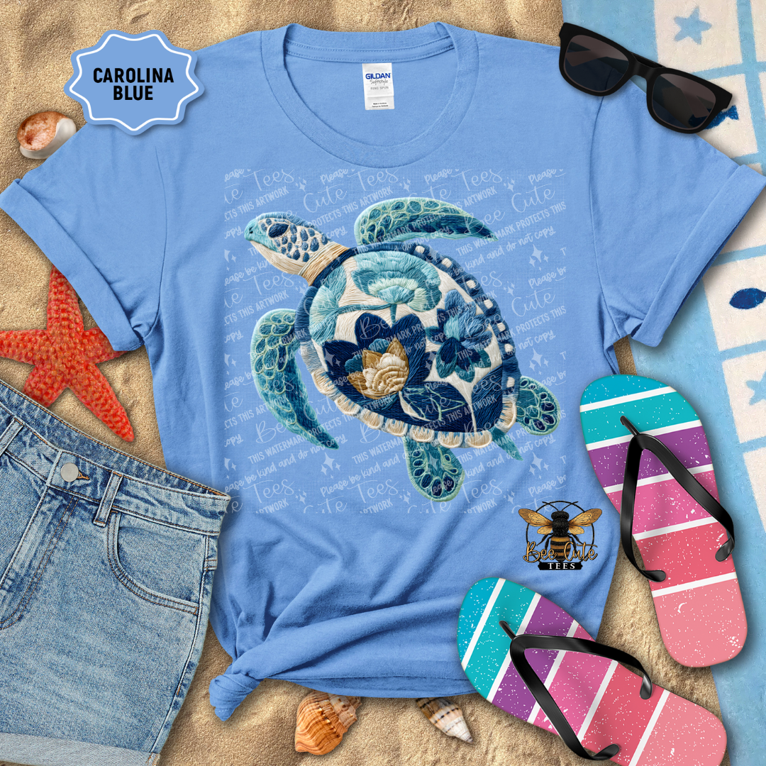 Blue t-shirt with a turtle design on a sandy surface with beach accessories.