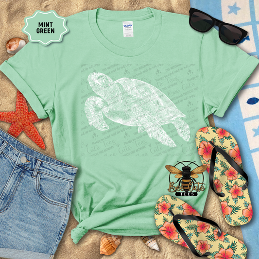 Mint green t-shirt with a turtle design on a beach-themed background with sunglasses, flip-flops, and starfish.