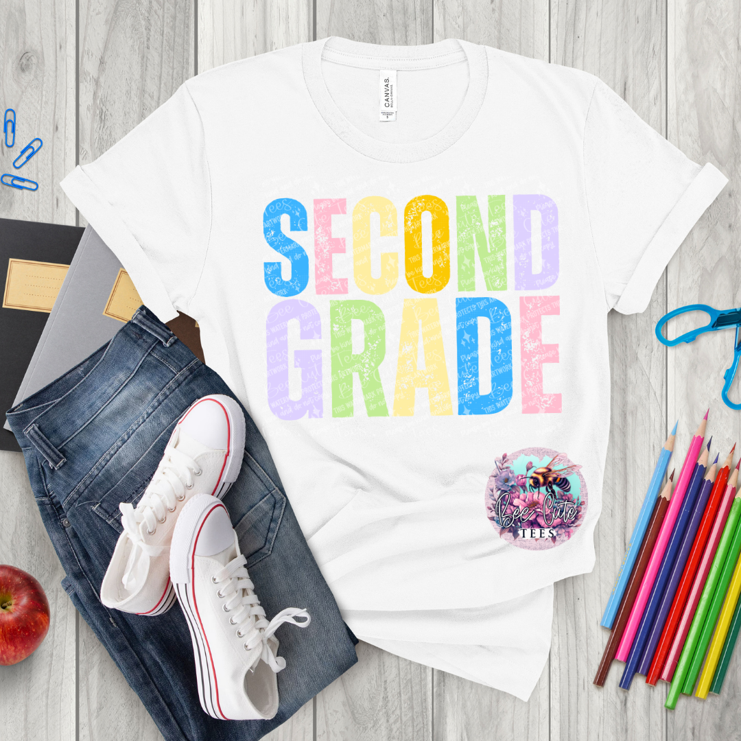 White t-shirt with 'SECOND GRADE' in colorful letters, jeans, sneakers, and school supplies on a wooden surface.
