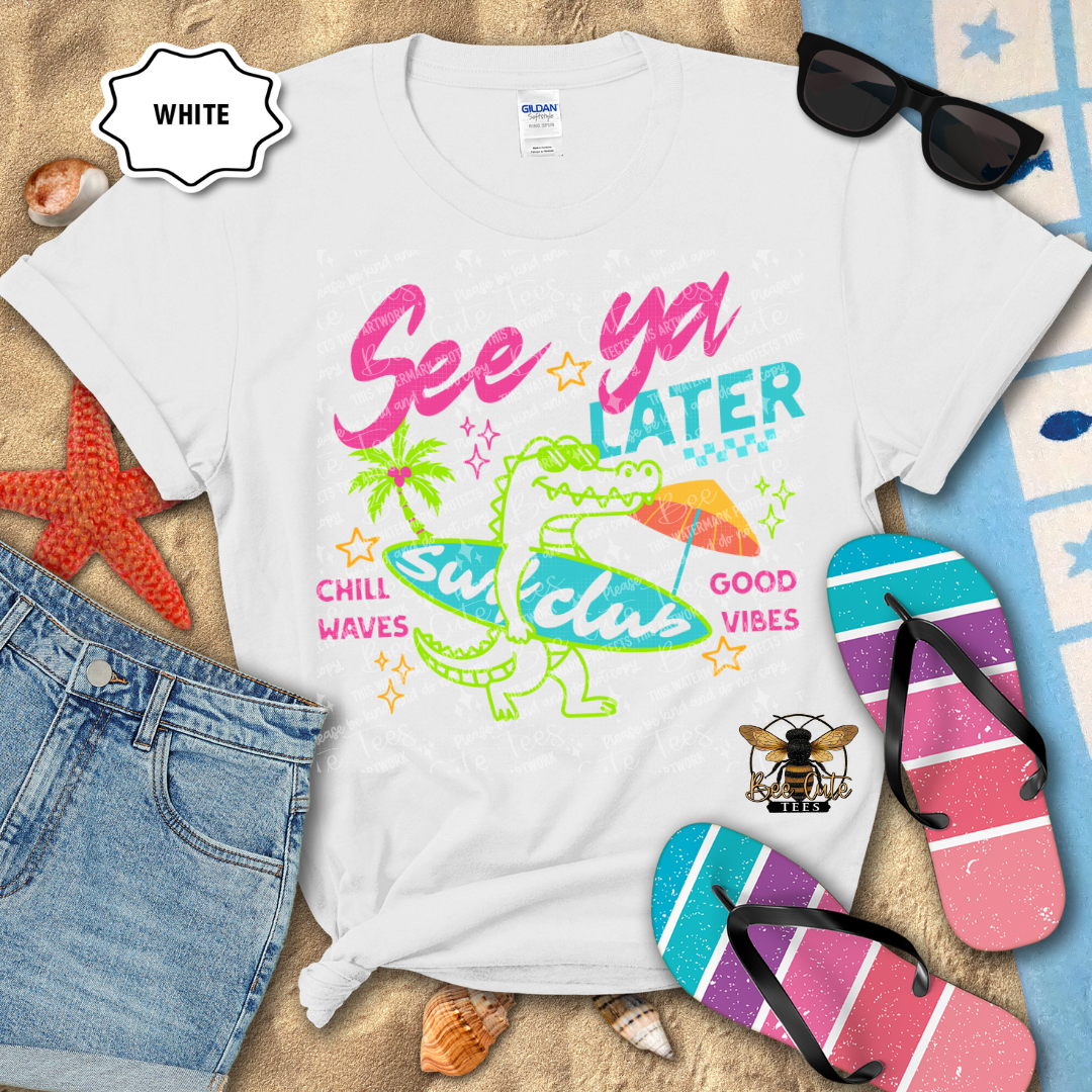 White t-shirt with colorful graphics and text, surrounded by beach accessories on a sandy background.