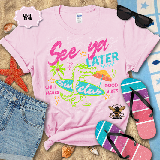 Light pink t-shirt with colorful graphics and text, surrounded by beach accessories on sand.