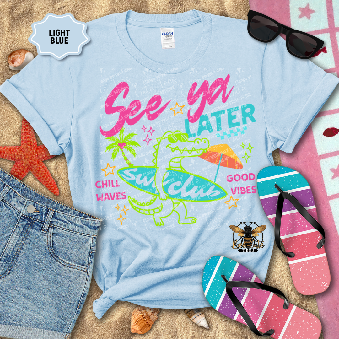 Light blue t-shirt with colorful graphics and text on a sandy background with beach accessories.