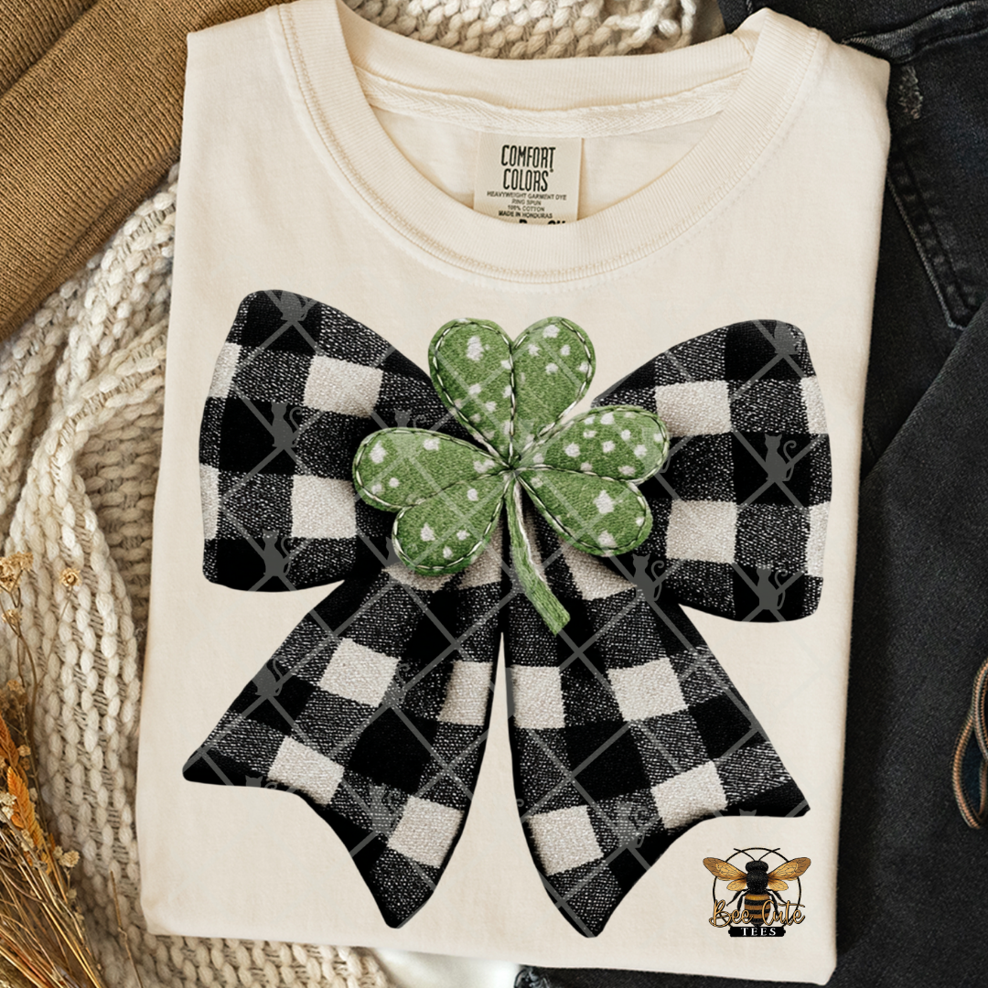 Folded white t-shirt with black and white checkered bow and green shamrock on a textured surface.
