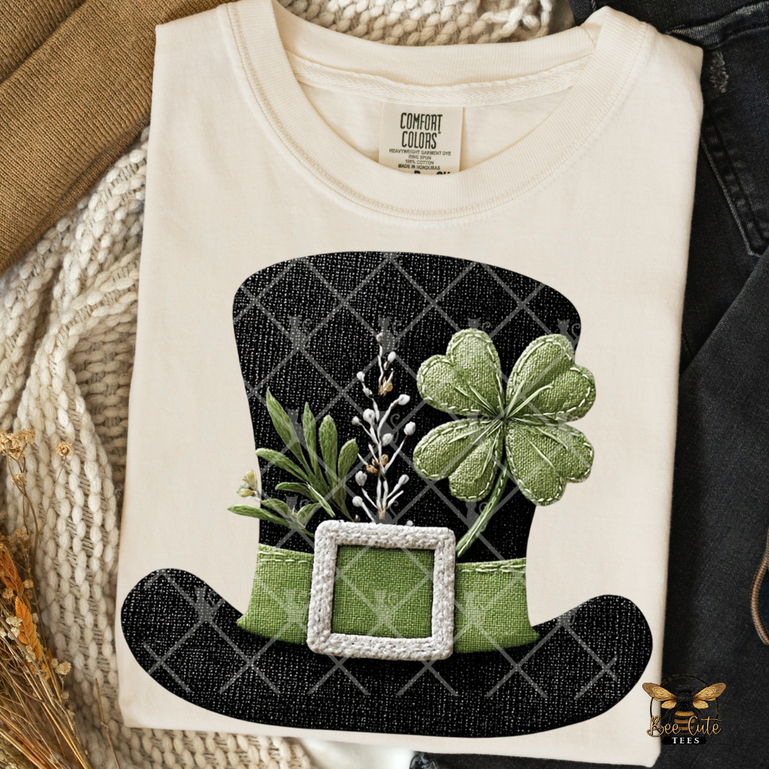 White t-shirt with a black hat and green shamrock design on a textured surface.