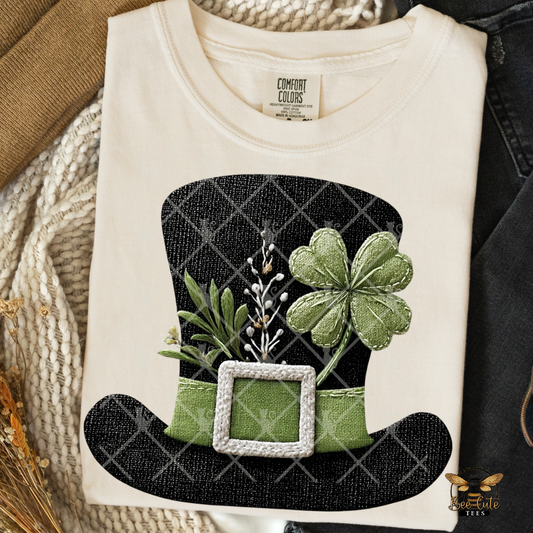 White t-shirt with a black hat and green shamrock design on a textured surface.