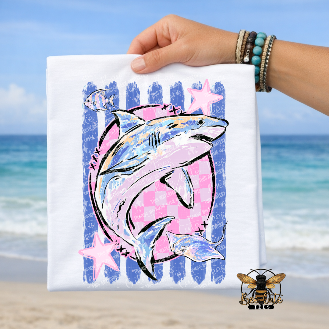 A hand holds a white t-shirt featuring a colorful graphic of a shark, fish, and stars on a beach background. The shirt has a pink and blue checkerboard pattern with blue vertical stripes and pink star accents.