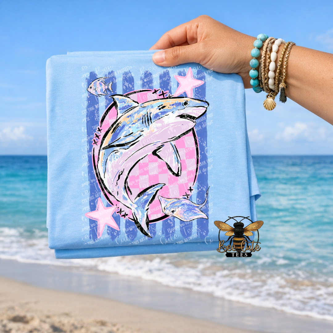 A folded light blue T-shirt is held up by a person's hand against a beach background. The shirt features a graphic print of a shark inside a pink circle with blue stripes and pink stars.