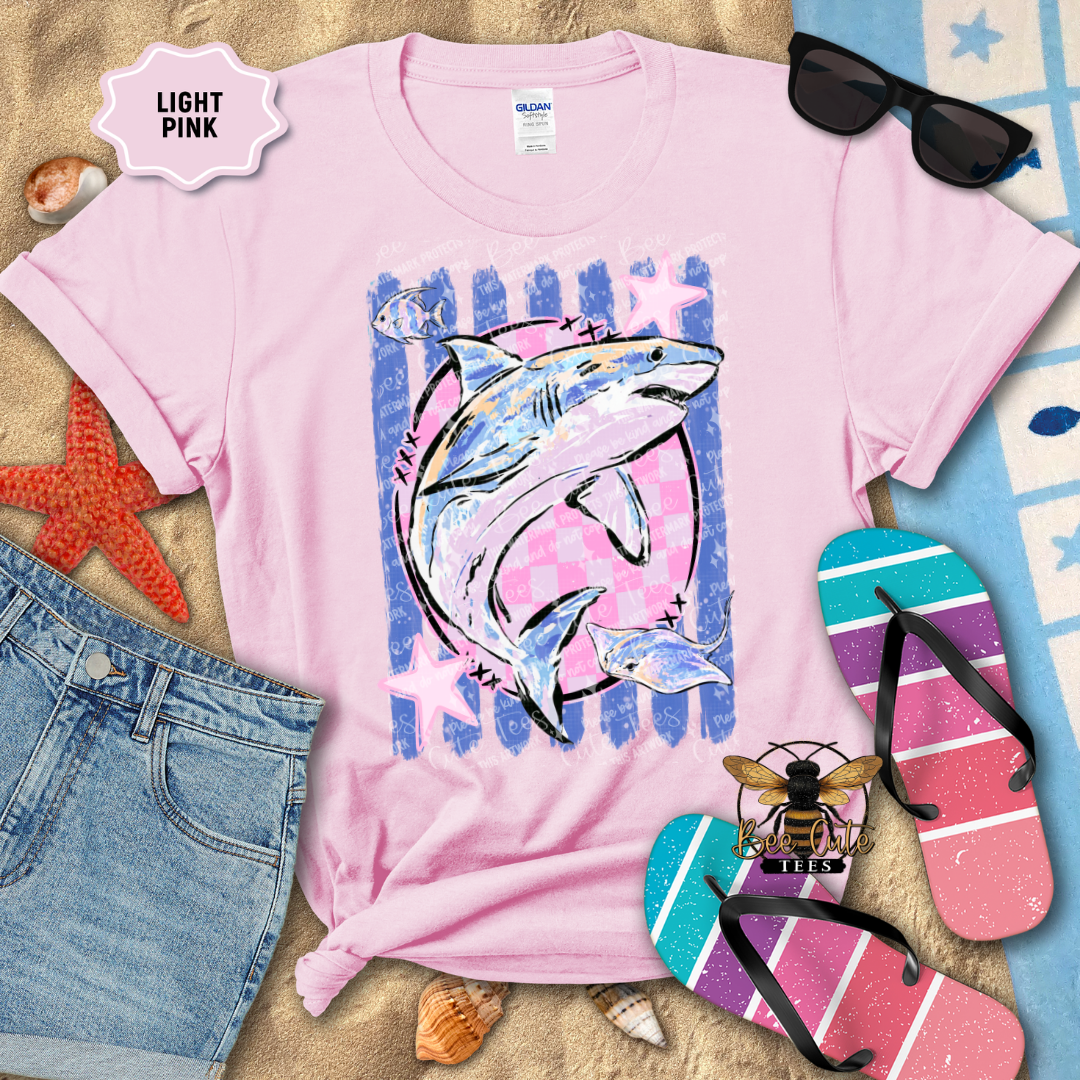 A light pink short-sleeve graphic T-shirt laid flat on a sandy surface with beach-themed props, including sunglasses, flip-flops, denim shorts, shells, and a starfish. The shirt has a large shark print in blue, white, and pink with the words 'LIGHT PINK' shown in a badge.