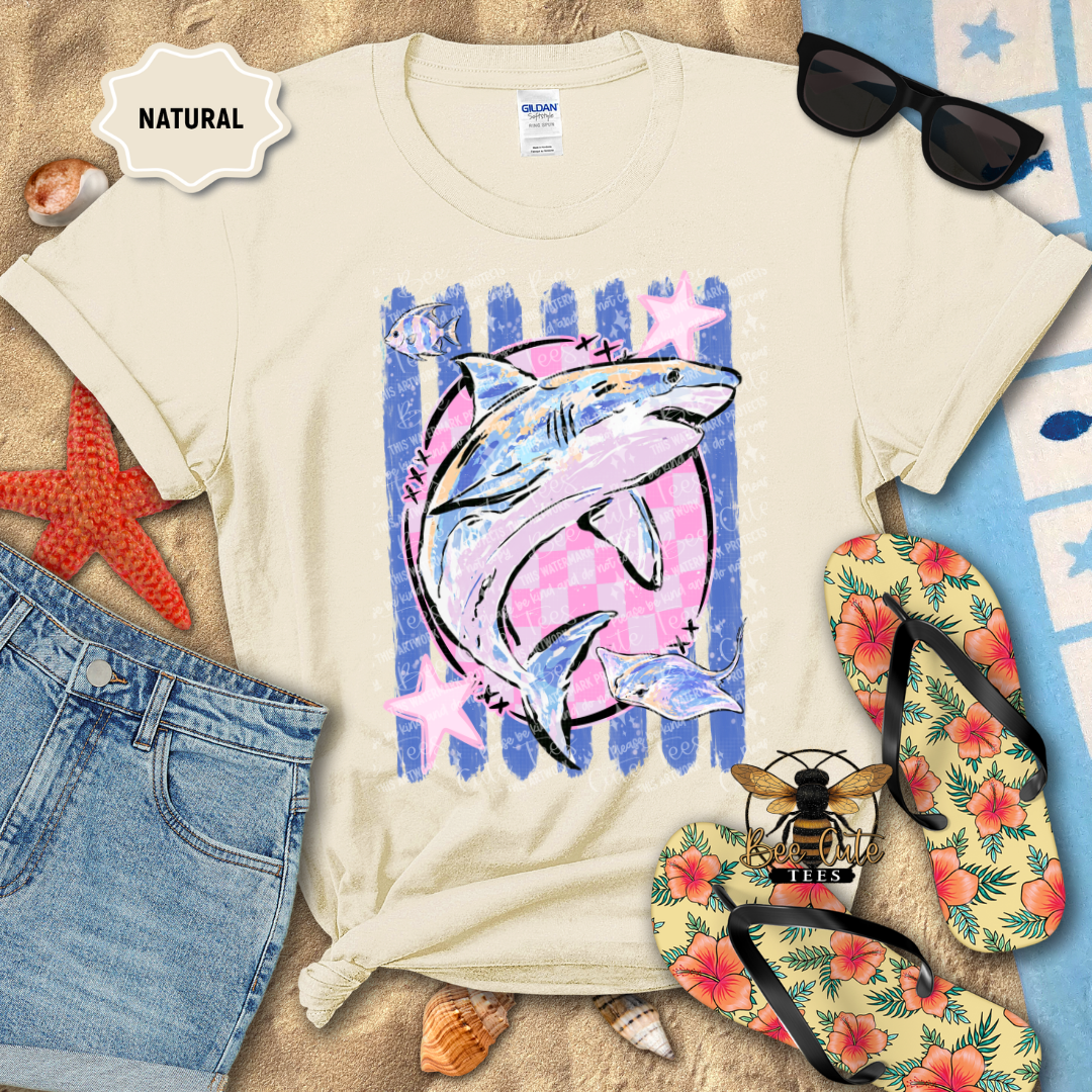 A cream-colored t-shirt with a graphic of a shark swimming in front of blue and pink striped background with pink stars. The t-shirt is styled in a flat-lay beach scene with blue denim shorts, yellow floral flip-flops, black sunglasses, a red starfish, and a bee logo, with a 'NATURAL' label sticker on the top left.
