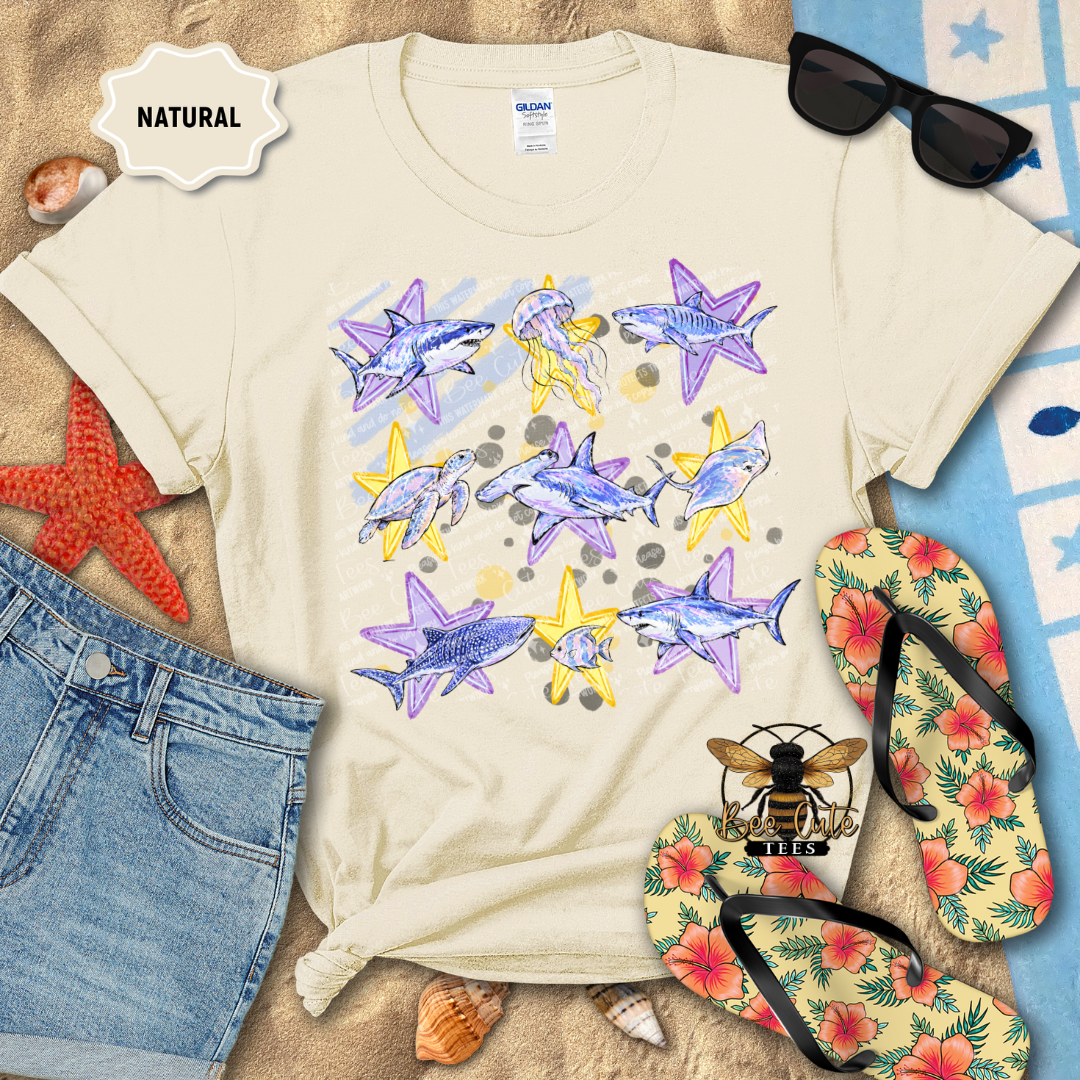 A cream-colored Gildan t-shirt with a graphic print of purple and yellow starfish, styled in a flat-lay beach-themed arrangement with denim shorts, sunglasses, tropical flip-flops, and seashells.
