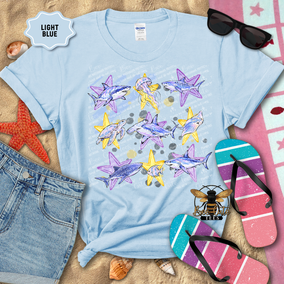 A light blue graphic t-shirt featuring a watercolor print of sharks and starfish, styled in a flat-lay arrangement with denim shorts, flip-flops, sunglasses, and seashells on a sandy background. A 'LIGHT BLUE' label is visible in the top left corner.