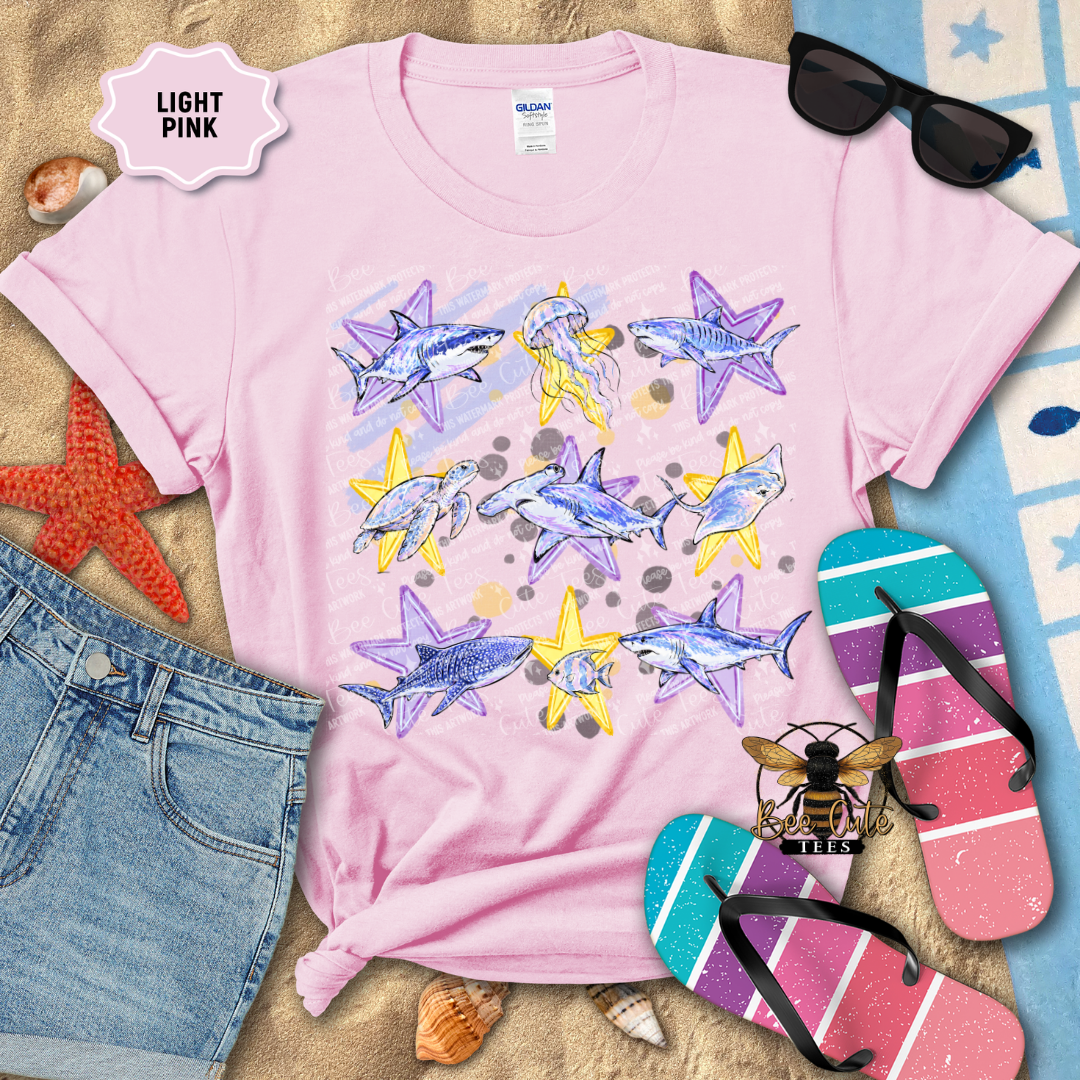 A light pink short-sleeve graphic T-shirt laid flat on a sandy surface with beach-themed props, including sunglasses, flip-flops, denim shorts, shells, and a starfish. The shirt has a large ocean-themed print with purple sharks, yellow starfish, jellyfish, and blue-gray starfish on a pale pink watercolor-style background.