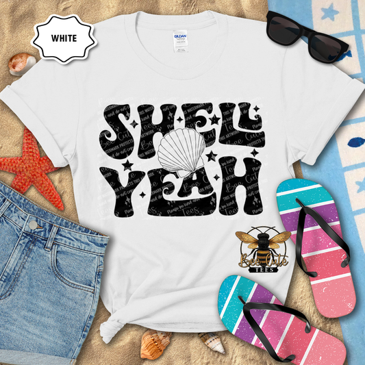 White t-shirt with 'Shell Yeah' design on a beach-themed background with flip-flops and sunglasses.