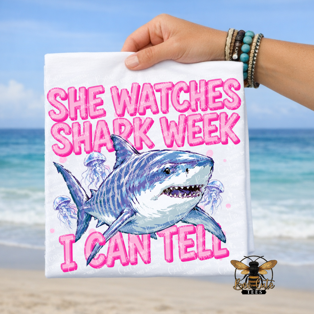 A hand holds a white t-shirt featuring a cartoon shark and the text 'SHE WATCHES SHARK WEEK' in pink, with a beach background.