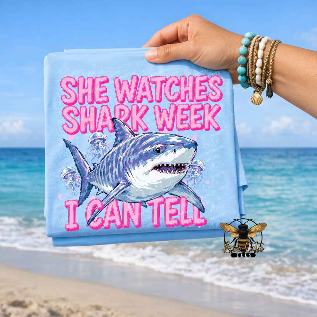 A folded light blue T-shirt is held up by a person's hand against a beach background. The shirt features a graphic of a blue and white shark with pink text reading 'SHE WATCHES SHARK WEEK'.