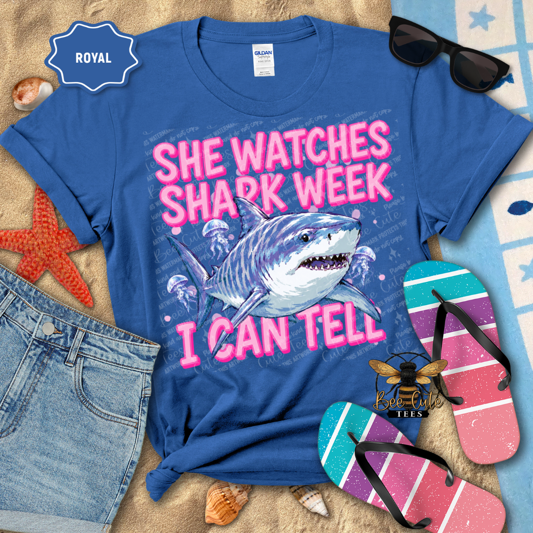 A royal blue graphic t-shirt featuring a shark illustration and the text 'SHE WATCHES SHARK WEEK' in pink. The shirt is styled in a flat-lay beach-themed arrangement with denim shorts, sunglasses, flip-flops, a starfish, and seashells on a sandy background.