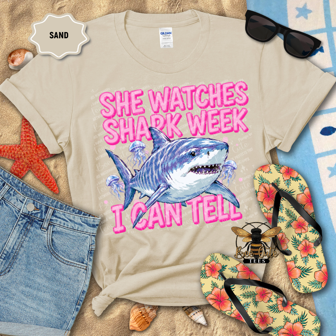 A beige graphic t-shirt laid flat on a sandy beach-themed background with sunglasses, denim shorts, tropical flip-flops, shells, and a starfish. The shirt features a blue shark illustration and the text 'SHE WATCHES SHARK WEEK' in pink.