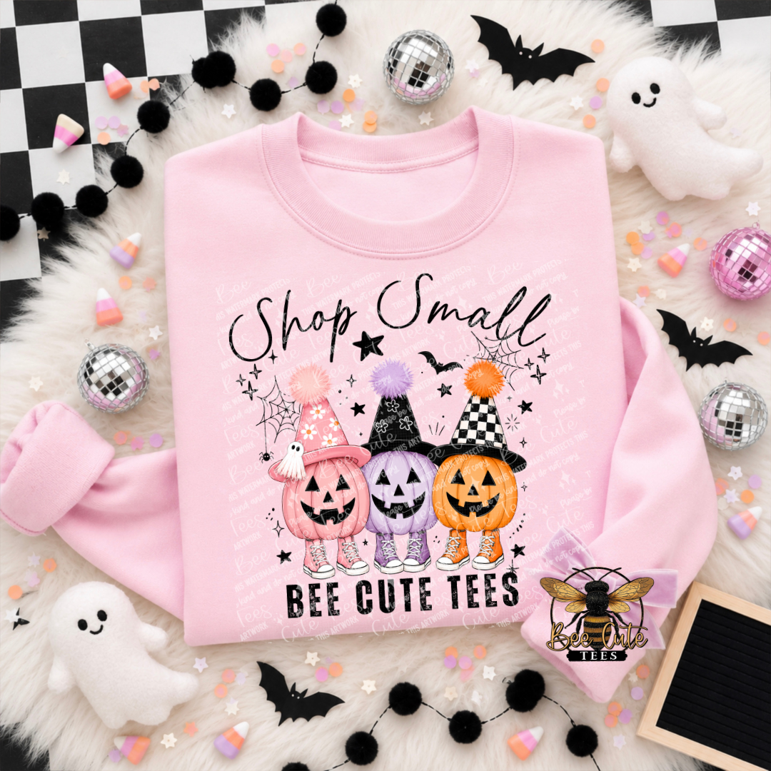 Folded light pink crewneck sweatshirt featuring a playful Halloween graphic with three smiling jack-o'-lanterns in pink, purple, and orange. Each pumpkin wears a festive witch hat and matching sneakers, surrounded by stars, bats, and spiderwebs. Black script reads “Shop Small” above the pumpkins, with “BEE CUTE TEES” printed below. Styled with pastel Halloween decorations.