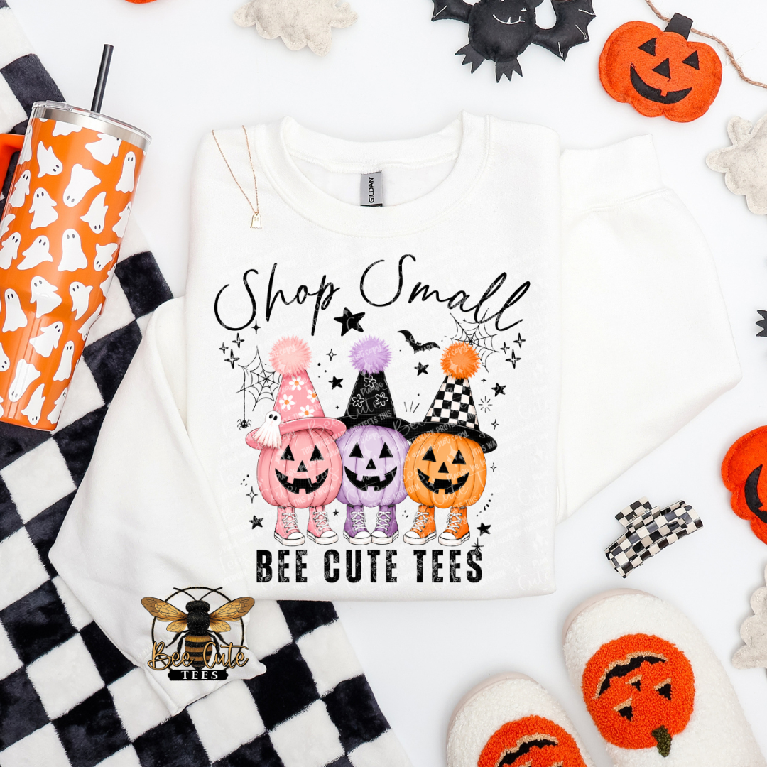 Folded white crewneck sweatshirt featuring three adorable jack-o'-lanterns in pink, purple, and orange, wearing patterned witch hats and colorful sneakers. Black “Shop Small” lettering appears above the pumpkins, with “BEE CUTE TEES” underneath. Surrounded by Halloween stars, bats, and spiderwebs, and styled with a ghost tumbler and festive accessories.