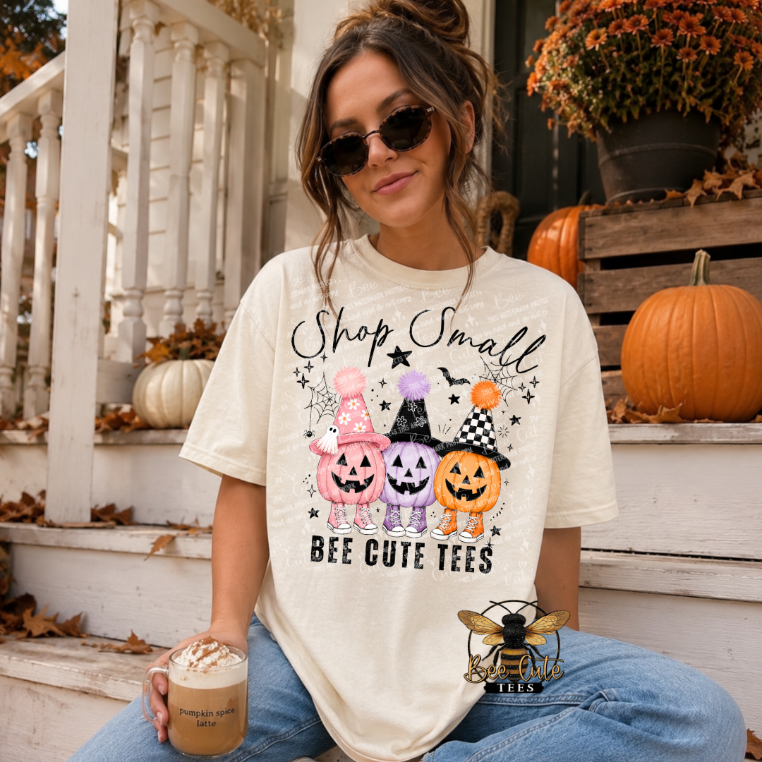 Woman wearing an oversized cream short-sleeve graphic tee featuring three colorful Halloween pumpkins dressed in witch hats and sneakers. The design reads “Shop Small” in black script above the pumpkins and “BEE CUTE TEES” below. Modeled with blue jeans and sunglasses while holding a pumpkin spice latte on a fall-decorated porch with pumpkins and autumn mums.