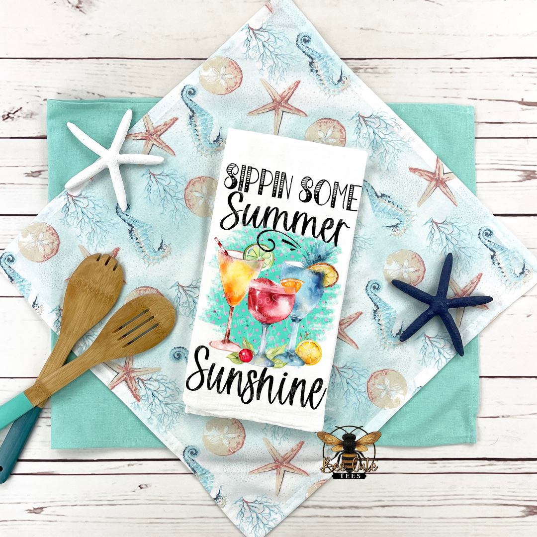 Summer-themed kitchen towel with cocktail design and text on a wooden surface with starfish and a spoon.