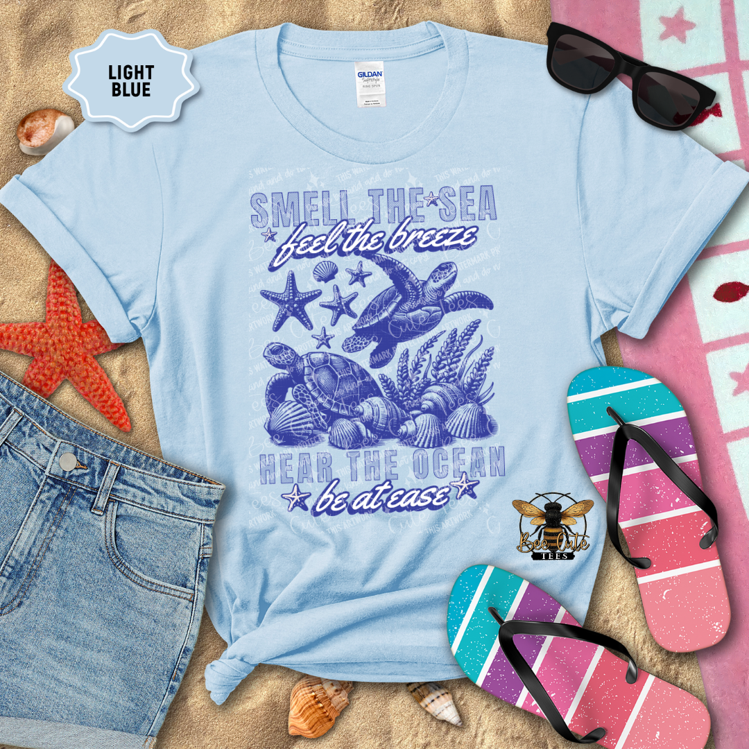 Light blue t-shirt with beach-themed design on a sandy background with flip-flops and sunglasses.