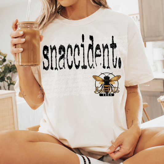 Person wearing a white t-shirt with 'snaccident' and a bee logo, holding a drink.