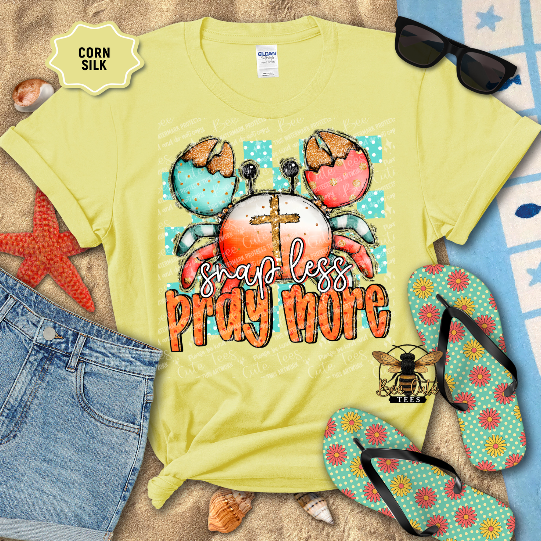 Yellow t-shirt with crab design and text 'Snap less, Pray more' on a beach setting with sunglasses and flip-flops.