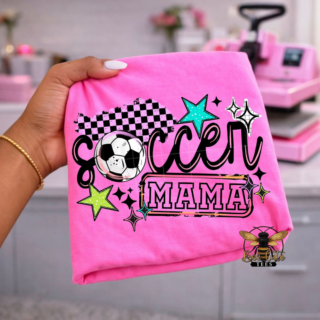 Pink t-shirt with 'Soccer Mama' design held by a hand, with a blurred background.