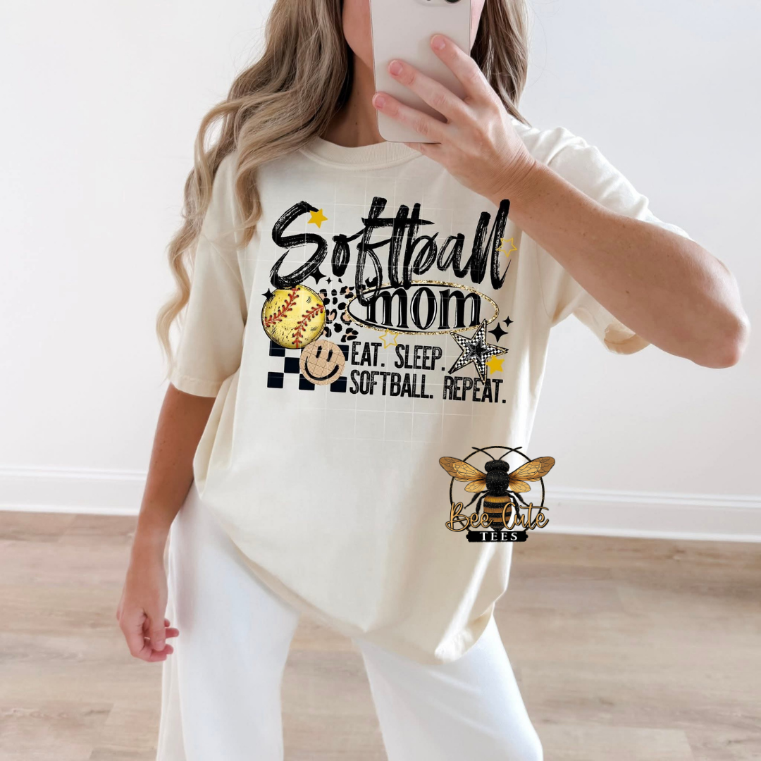 A woman takes a mirror selfie wearing a cream-colored graphic t-shirt with 'Softball Mom' written on the front, paired with white pants. 