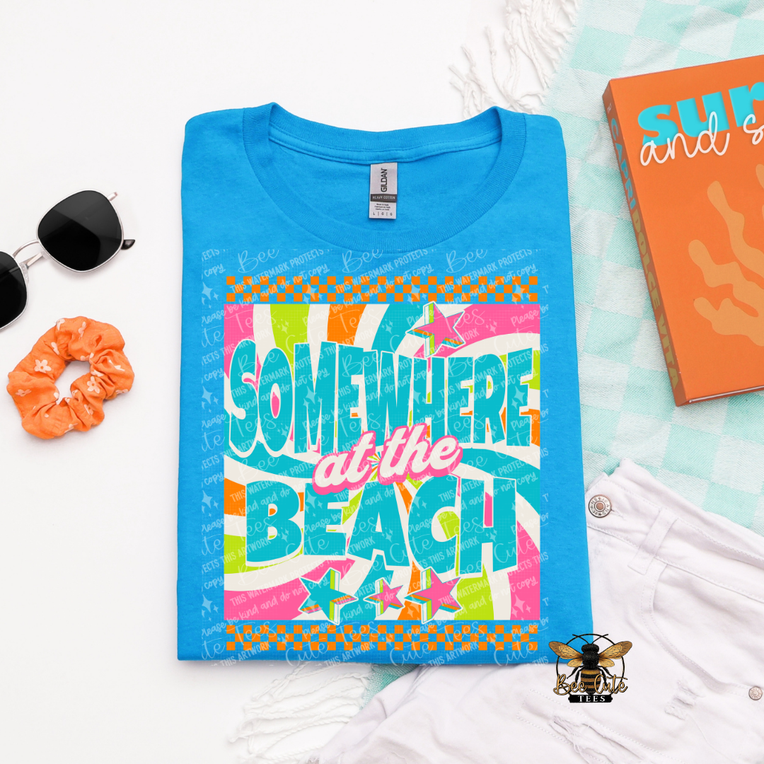 Blue t-shirt with 'Somewhere at the Beach' design on a white surface with accessories.