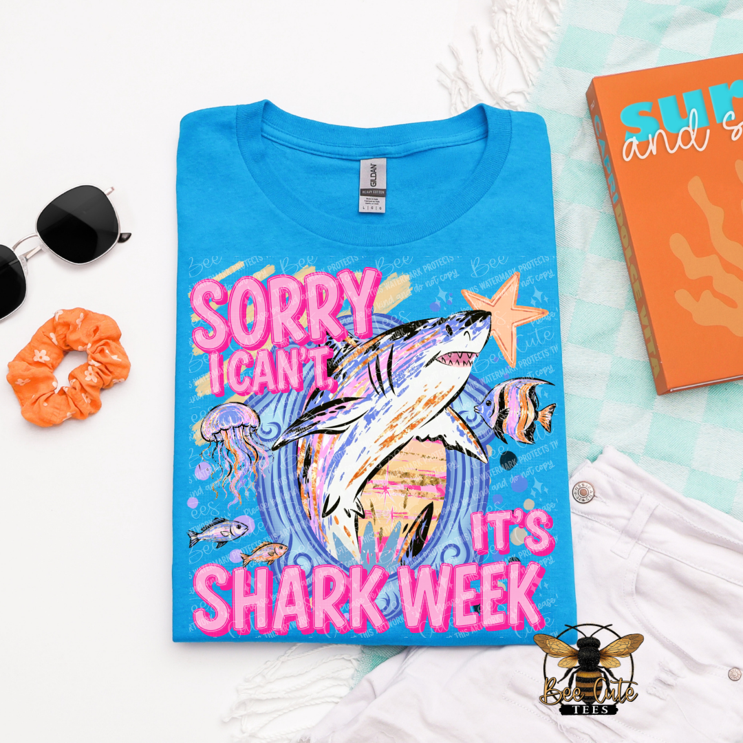 A folded bright blue graphic T-shirt is displayed in a styled flat lay on a white surface. The shirt features a colorful shark illustration with the text 'SORRY I CAN'T IT'S SHARK WEEK' in pink and orange.
