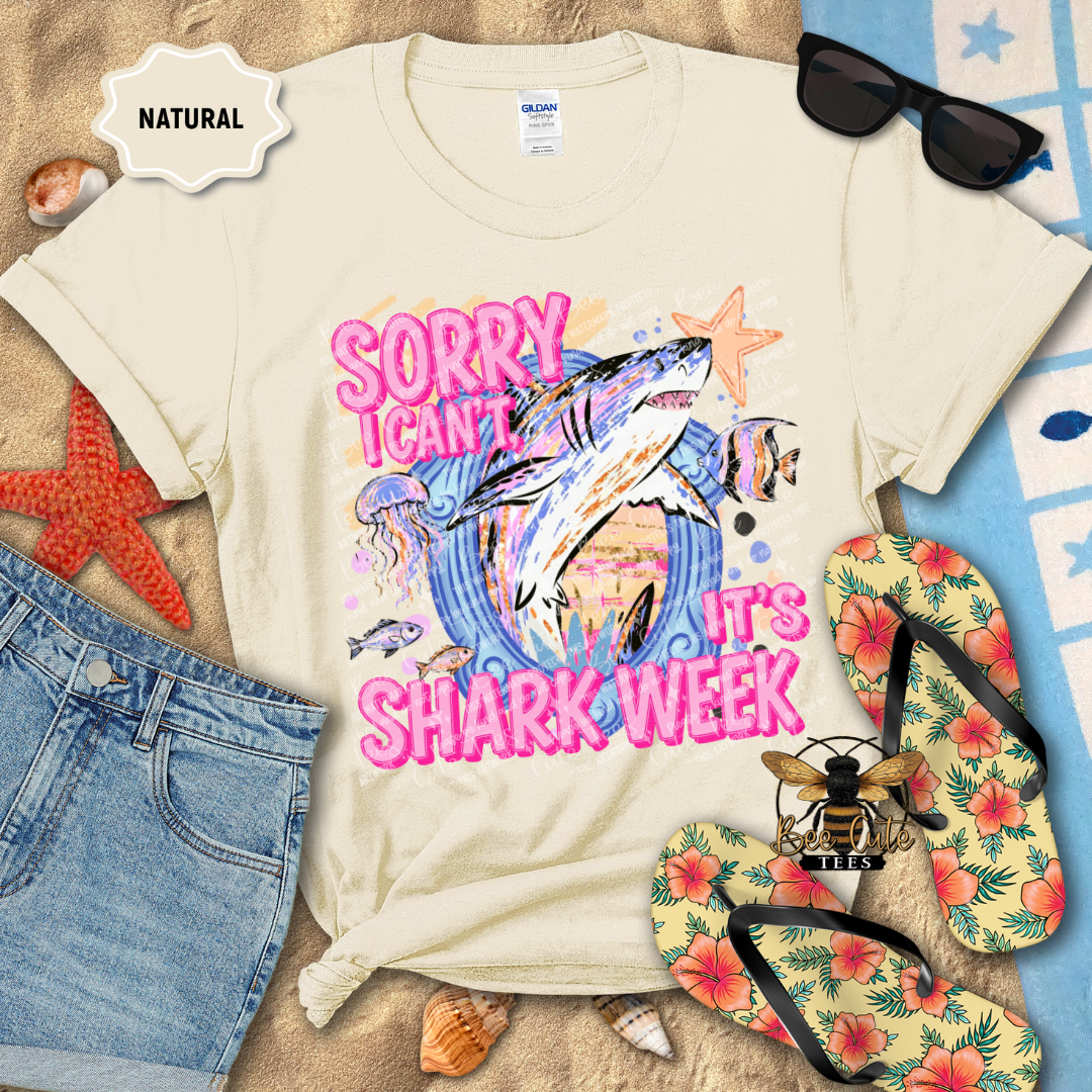 A flat-lay of a cream-colored Gildan t-shirt with a colorful shark graphic and the text 'SORRY I CAN'T SHARK WEEK' in pink. The shirt is styled with blue denim shorts, tropical flip-flops, sunglasses, starfish, and seashells on a sandy beach-themed background.