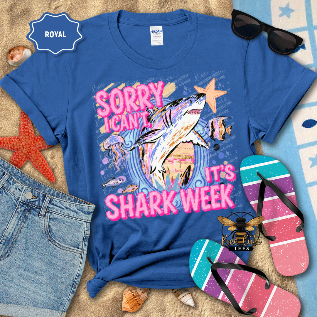A royal blue Gildan t-shirt with a colorful shark graphic and the text 'Sorry I Can't It's Shark Week' in pink and white. The shirt is styled in a flat lay with denim shorts, sunglasses, flip-flops, and starfish on a sandy beach-themed background.