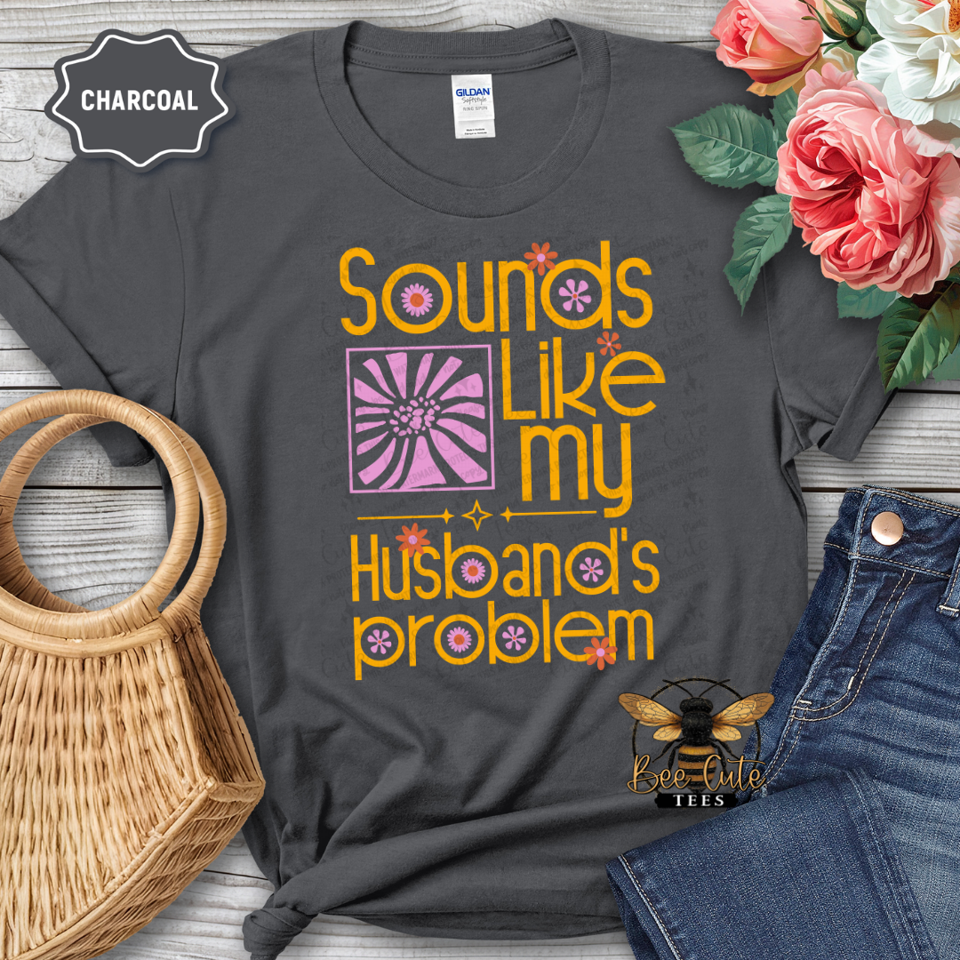 Gray t-shirt with 'Sounds like my husband's problem' text, surrounded by flowers and a handbag on a wooden surface.