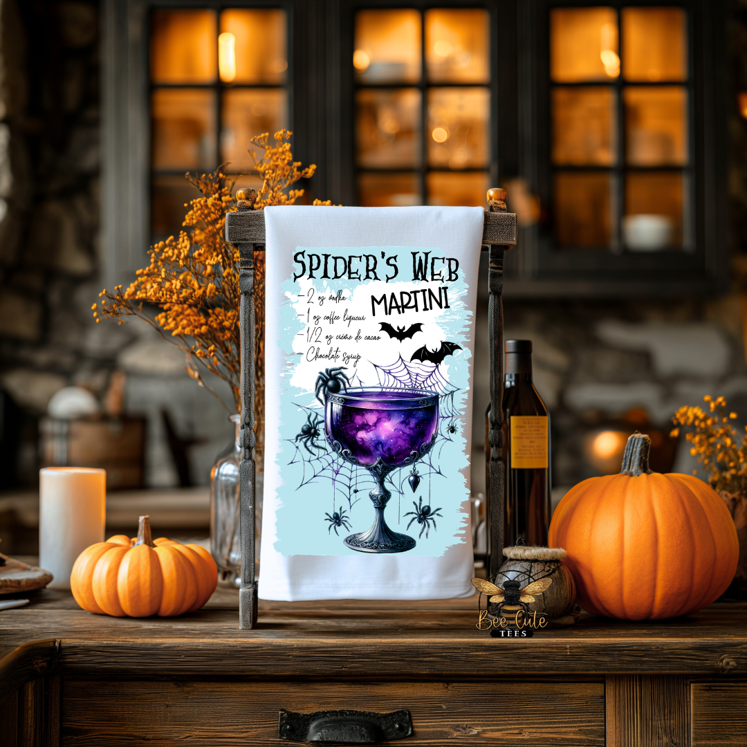 A white tea towel with a Halloween-themed 'Spider's Web Martini' graphic is displayed on a rustic wooden stand. The scene is styled as a cozy autumn kitchen or bar counter with pumpkins, candles, and a bottle of wine, creating a warm, festive atmosphere.