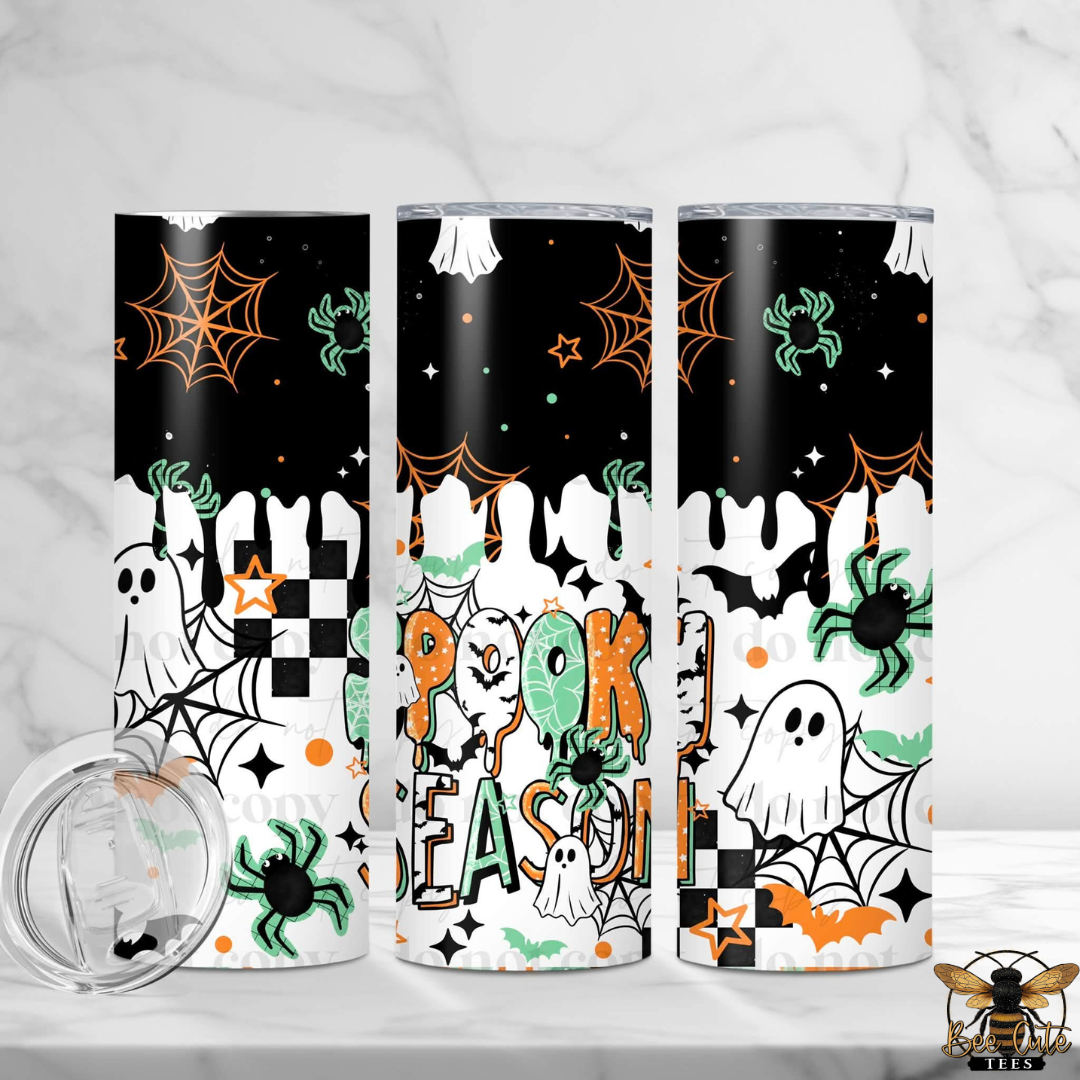 Three slim tumblers with a Halloween-themed print are displayed on a white marble surface. The design features a black and white dripping pattern with orange spiderwebs, green spiders, ghosts, bats, stars, and the words 'Halloween' in orange and green.