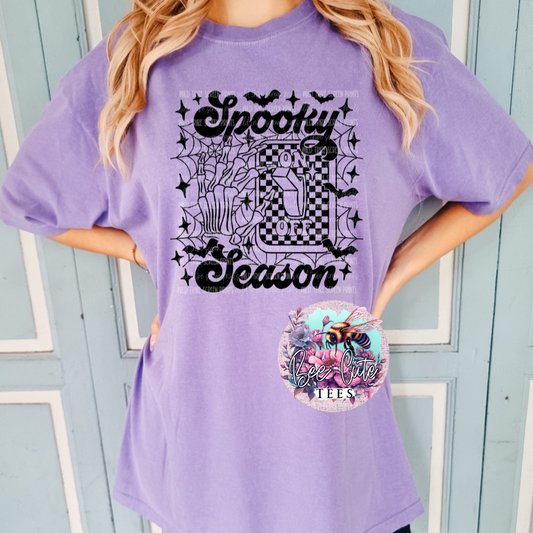 Purple Halloween graphic tee featuring “Spooky Season” lettering with skeleton hands flipping an “On/Off” switch to on, surrounded by spiderwebs, bats, stars, and checkerboard details.
