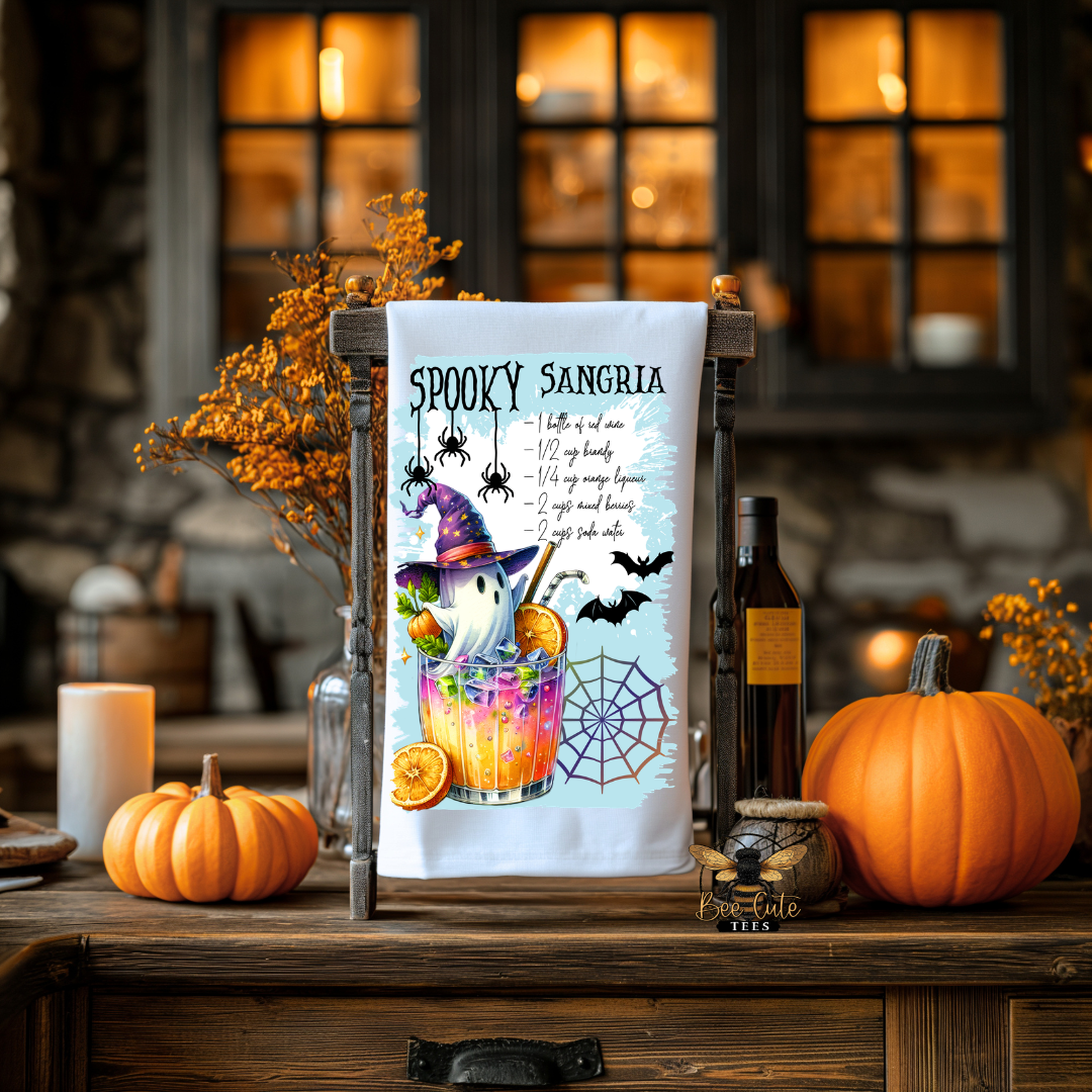 A white kitchen tea towel with a colorful 'Spooky Sangria' recipe print, displayed on a wooden stand on a rustic wooden table surrounded by pumpkins, candles, and autumn decorations.