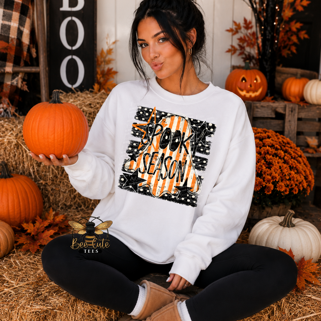 A woman sitting cross-legged on a hay bale in a fall-themed setting, wearing a white crewneck sweatshirt with a 'Halloween Season' graphic. She is holding a pumpkin, with other pumpkins, orange flowers, and a rustic wooden background behind her.