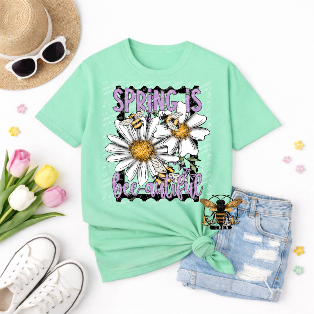 Mint green t-shirt with 'Spring is bee-utiful' design, surrounded by flowers, sunglasses, and a hat on a white background.